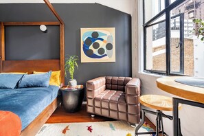 A modern bedroom with a bed, a cushioned seating area, a small table, and a window with a view of buildings. - Cosy CBD Loft Walk to Dining+Shopping Zodiak Stays (Auckland)