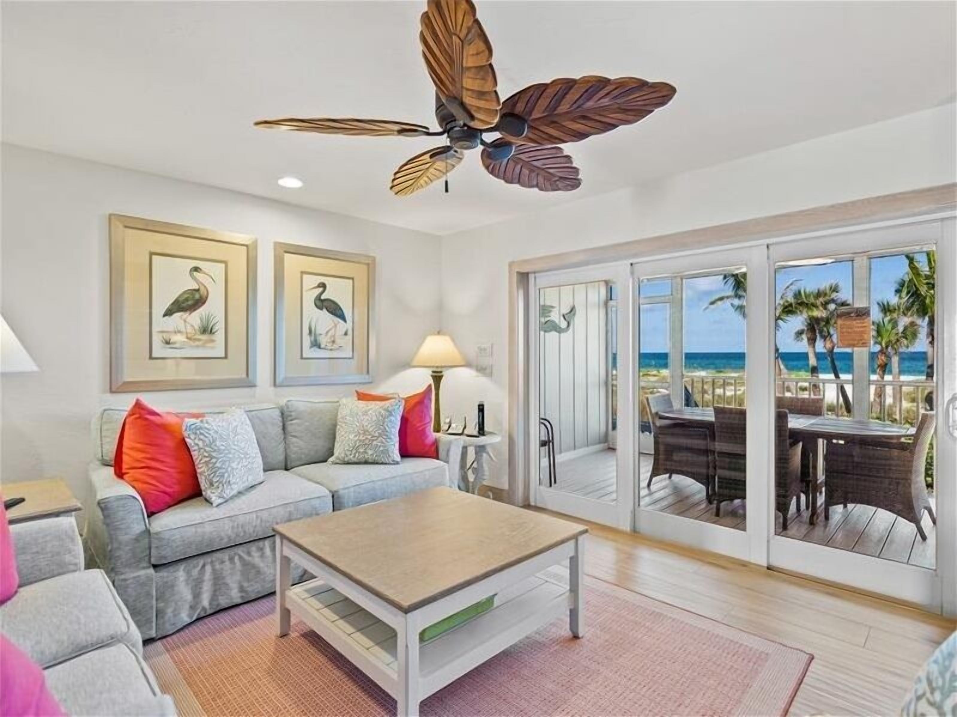 A living room with a grey sofa, a wooden coffee table, and a ceiling fan. There are framed pictures on the wall, a lamp, and a view of the beach through large sliding doors.