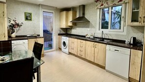 Une cuisine avec des meubles en bois, un lave-linge, un lave-vaisselle et un coin repas comprenant une table et des chaises en verre.