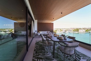 Uma área moderna de refeições ao ar livre com vista para a água, mobiliada com sofás brancos e uma mesa de madeira.