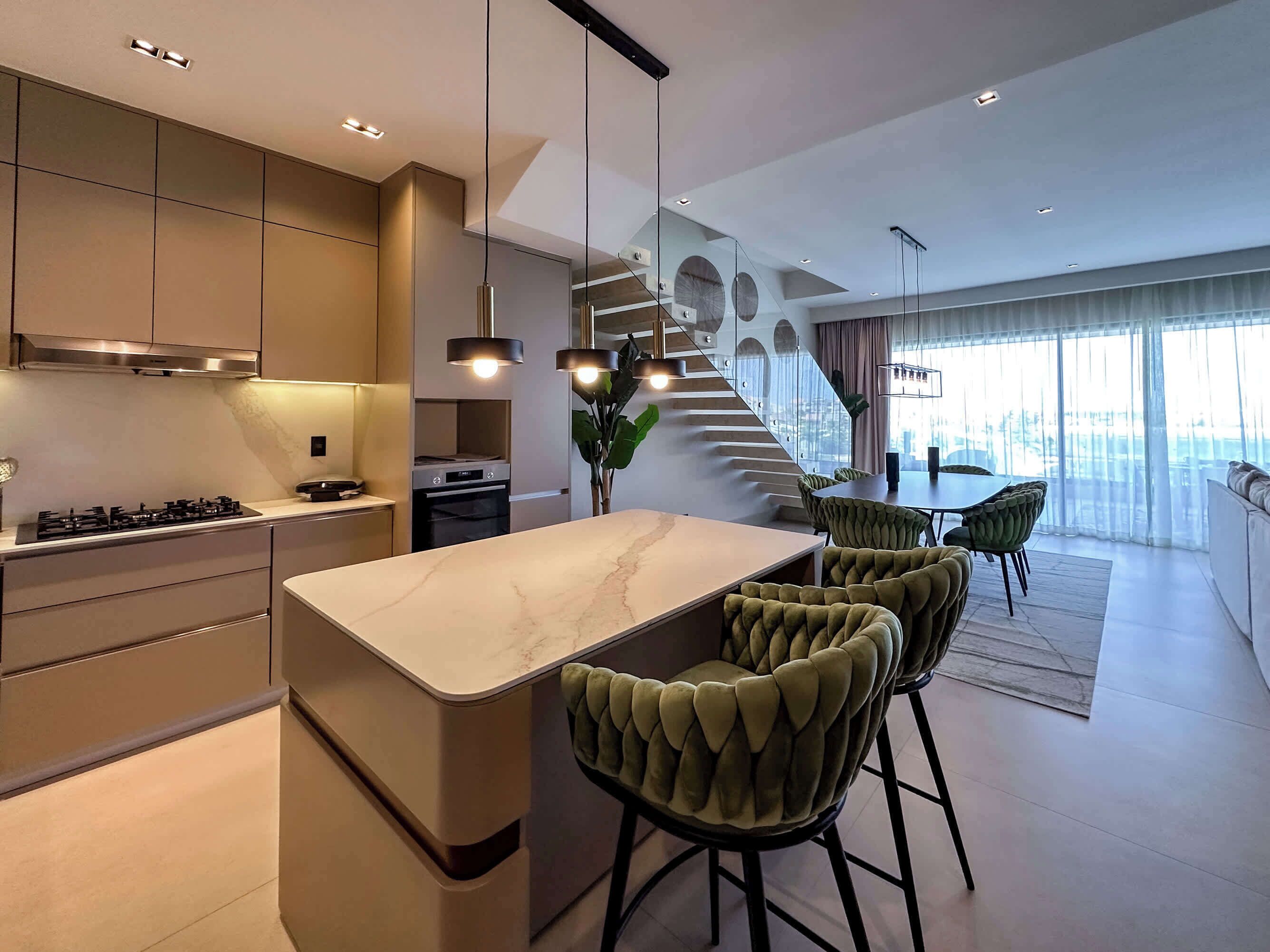 Une cuisine moderne dotée d’un îlot central, d’appareils électroménagers intégrés et d’un escalier menant à un coin repas avec des chaises recouvertes d’un tissu vert.