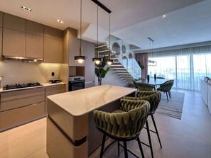 A modern kitchen with a central island, built-in appliances, and a staircase leading to a dining area with green upholstered chairs. - Spectacular Marina Penthouse w private rooftop (Punta Cana)