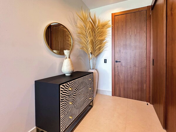 Un couloir avec une console noire, un miroir rond, un vase contenant des plantes séchées et une porte en bois.