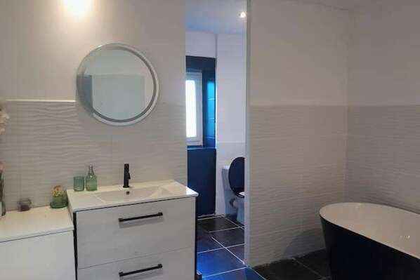 Un baño moderno con tocador blanco, espejo redondo, bañera negra y un inodoro visible al fondo.