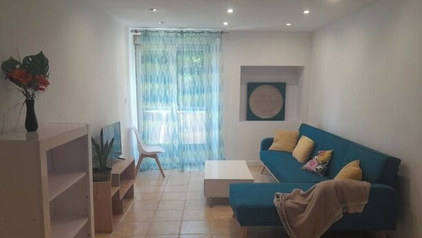 A living room with a blue sofa, a white coffee table, and a TV stand. There is a window with curtains, a plant on the left, and a wall-mounted artwork. - Stay in béarnais country (Baigts-de-Béarn)