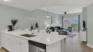 Cocina moderna con una isla grande, armarios blancos y lavavajillas. Hay una zona de comedor con sofá y mesa de centro. La habitación tiene un ventanal con vistas a edificios y vegetación.