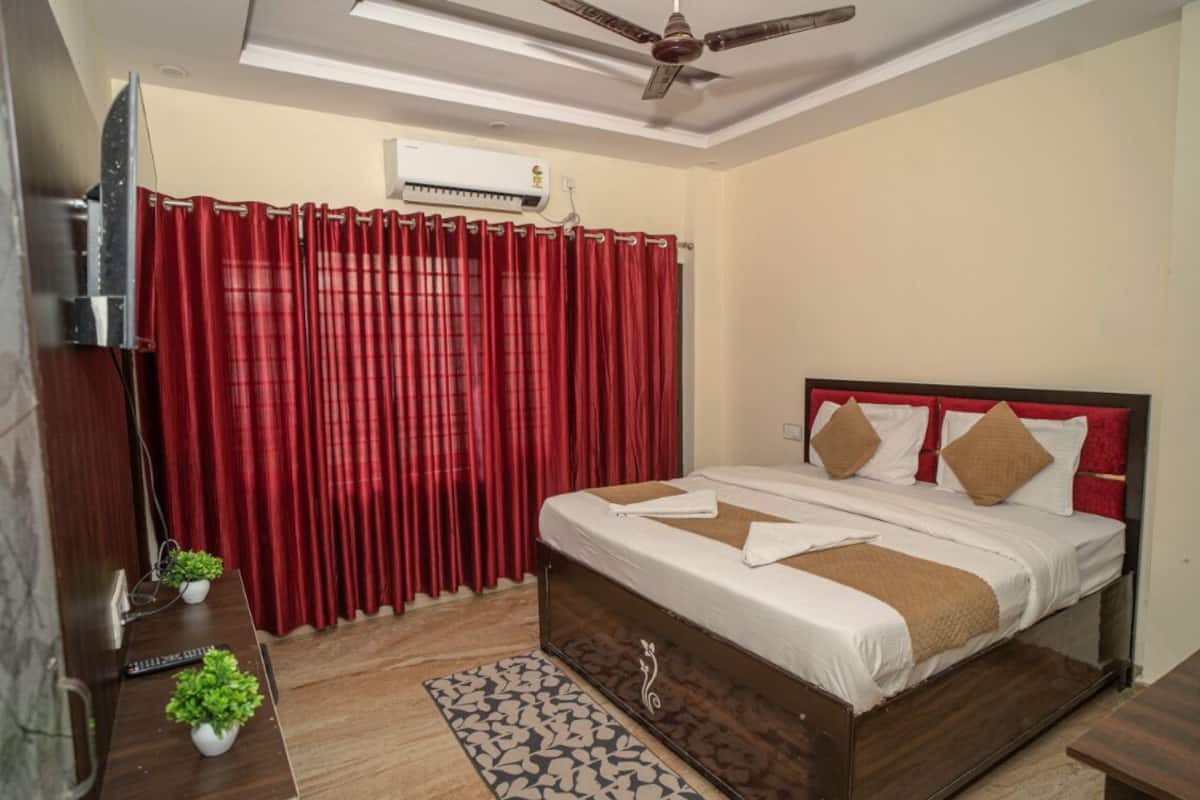 A bedroom with a bed, red curtains, a ceiling fan, an air conditioner, and a small table with plants.