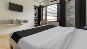 Une chambre d’hôtel avec un lit, un téléviseur fixé au mur, une fenêtre avec des stores, une chaise et une petite étagère.