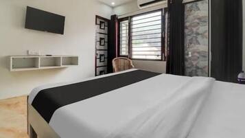A hotel room with a bed, a TV mounted on the wall, a window with blinds, a chair, and a small shelf.