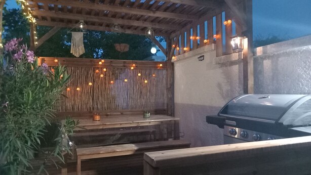 An outdoor patio with a wooden pergola, string lights, a barbecue grill, and a seating area. - Maison la Tourterelle in Auvergne (Champs)