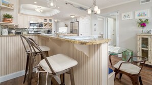 Una cocina con isla central, taburetes de bar, luces colgantes y un refrigerador.
