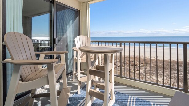 Un balcón con dos sillas Adirondack y una pequeña mesa, con vista a una playa y una vista despejada del océano.