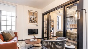 Ein modernes Wohnzimmer mit Ledersofa, Couchtisch und gemustertem Sessel. An der Wand ist ein Flachbildfernseher angebracht, und ein großes Fenster bietet Blick auf ein Schlafzimmer.