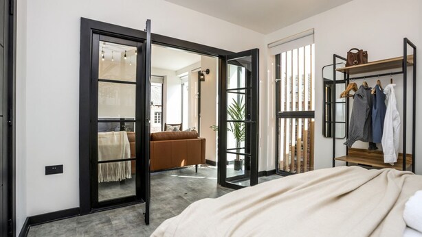 A modern bedroom with a large bed, a wardrobe, and a sliding door leading to a living area with a sofa and a plant.