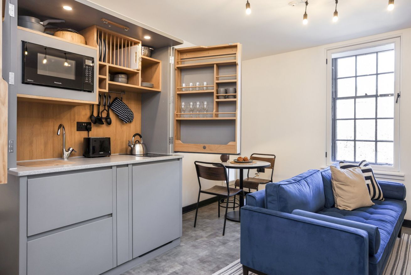 A compact kitchen with a microwave, kettle, and utensils on the counter. There is a blue sofa and a dining table with chairs.