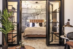 A modern bedroom with a large bed, a desk, and a chair. There is a plant on the left side and a lamp on the right side. - Locker Room:  Stylish 2bed w/City Views-3rd floor (Edinburgh)