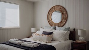 Un lit bien fait, recouvert d’une couverture bleu foncé et agrémenté d’oreillers blancs, un miroir rond accroché au mur et une table de chevet avec une lampe.