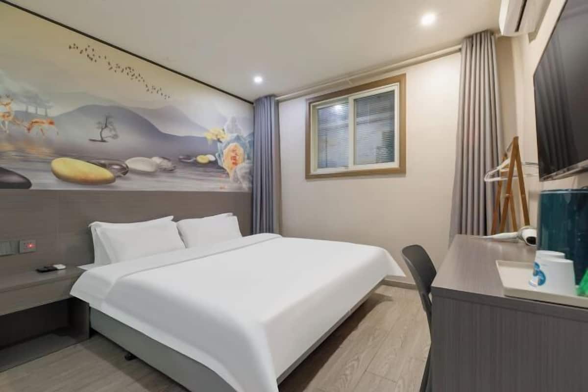 A hotel room with a bed, a desk with a chair, a window with blinds, and a wall with a mural.