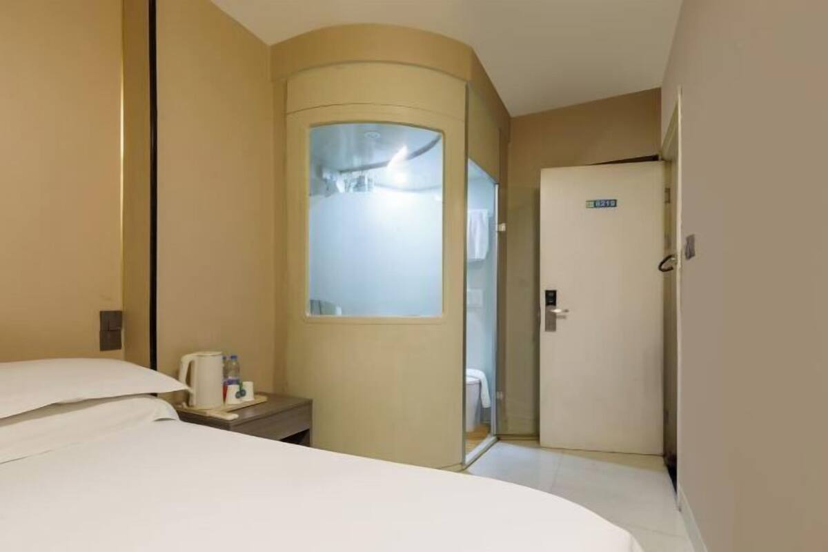 A hotel room with a bed, a bedside table with a coffee maker, a glass-enclosed shower, and a door with a sign.