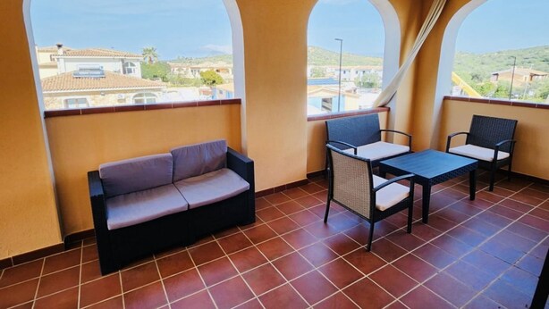 Un balcón con sofá y dos sillas, suelo de baldosas y amplias ventanas de arco que ofrecen vistas a edificios y colinas.