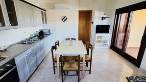 Una cocina compacta con muebles blancos, una mesa de comedor con sillas, un televisor y un ventanal corredero que da a un espacio exterior.