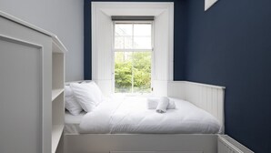 A bedroom with a single bed, white bedding, a window with a view of greenery, and a dark blue wall. - Haymarket - Single bunk room (Edinburgh)