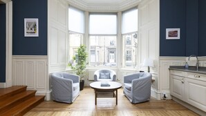 A room with a bay window, a round coffee table, two armchairs, a potted plant, and a view of buildings outside.
