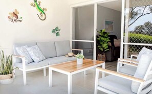Una sala de estar moderna con un sofá gris, sillones blancos, una mesa de centro de madera y un amplio ventanal corredizo que da a un espacio exterior.