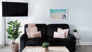 A modern living room with a black sofa, a flat-screen TV, a framed beach painting, and two potted plants.