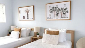 A hotel room with two beds, a wooden headboard, a small table with a lamp, and framed pictures on the wall.