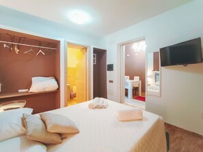 Habitación de hotel con una cama, un armario, un televisor y un baño visible a través de una puerta abierta.