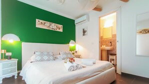 A bedroom with a green accent wall, a white bed with patterned pillows, a nightstand with a lamp, and a bathroom visible through an open door. - villa di l'abbiu country relax (Sorso)