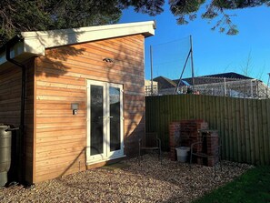 A wooden building with a sliding glass door, a metal chair, and a brick structure in the background. - Y Caban-the Cabin-1 Bed-sleeps 2-parking-fast Wifi (Llanelli)