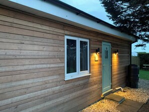 A wooden building with a window and a door, a small outdoor area with gravel, and a black barrel. - Y Caban-the Cabin-1 Bed-sleeps 2-parking-fast Wifi (Llanelli)