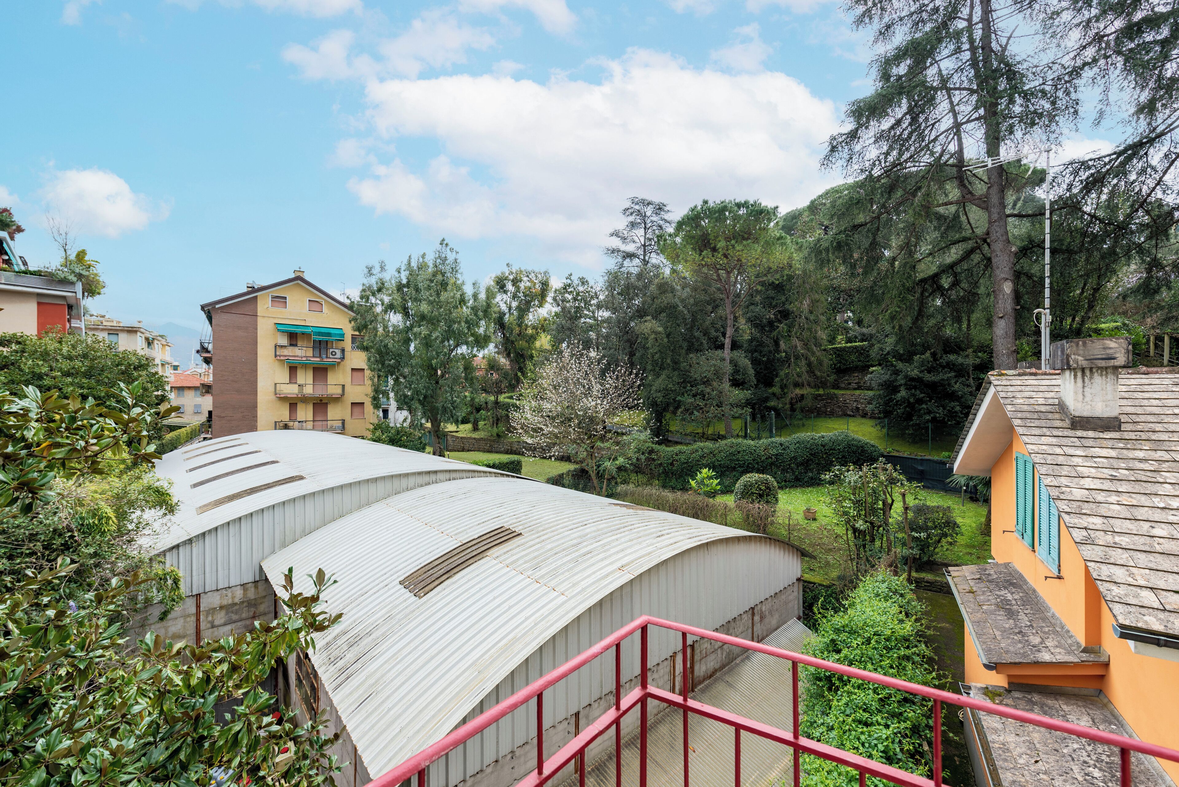 A residential area with a curved metal roof structure, a yellow building with a balcony, and a green landscape with trees and a small water channel.