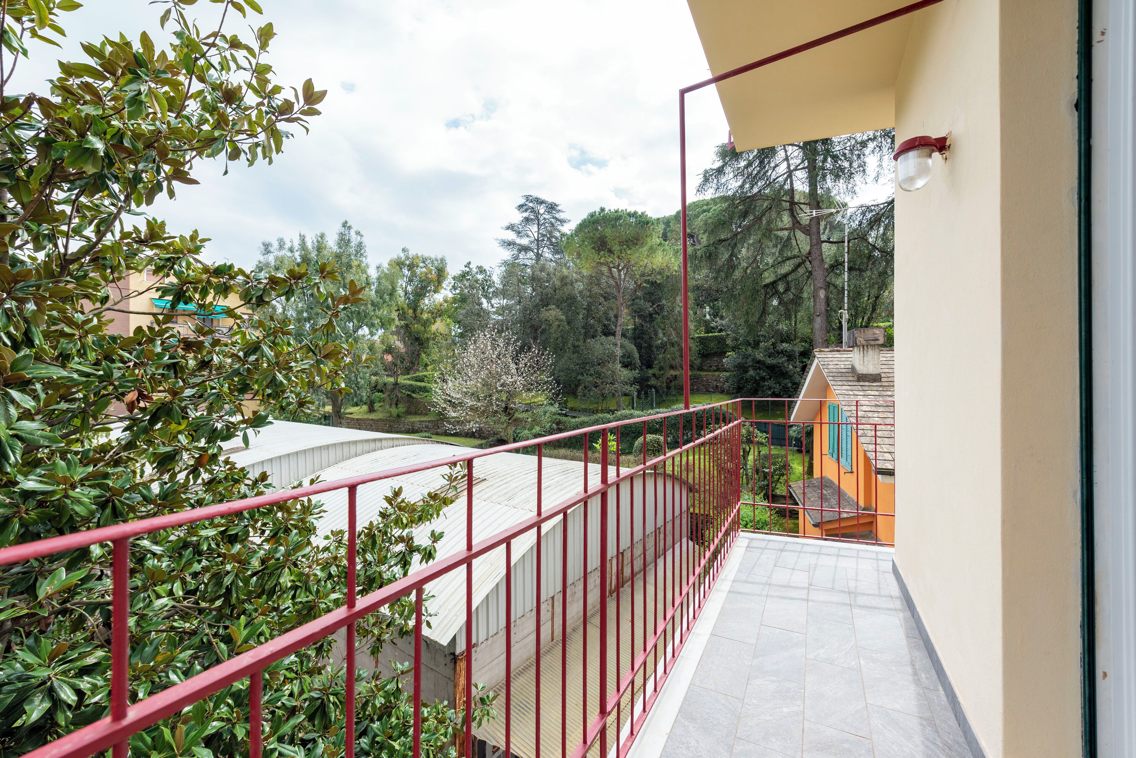 Un balcón con barandilla roja que da a un jardín y árboles.