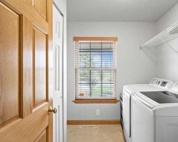 A laundry room with a washer and dryer, a window with blinds, and a wooden door.