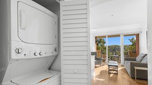 A modern laundry room with a built-in washer and dryer, a sliding door leading to a balcony with outdoor furniture and a view of greenery.