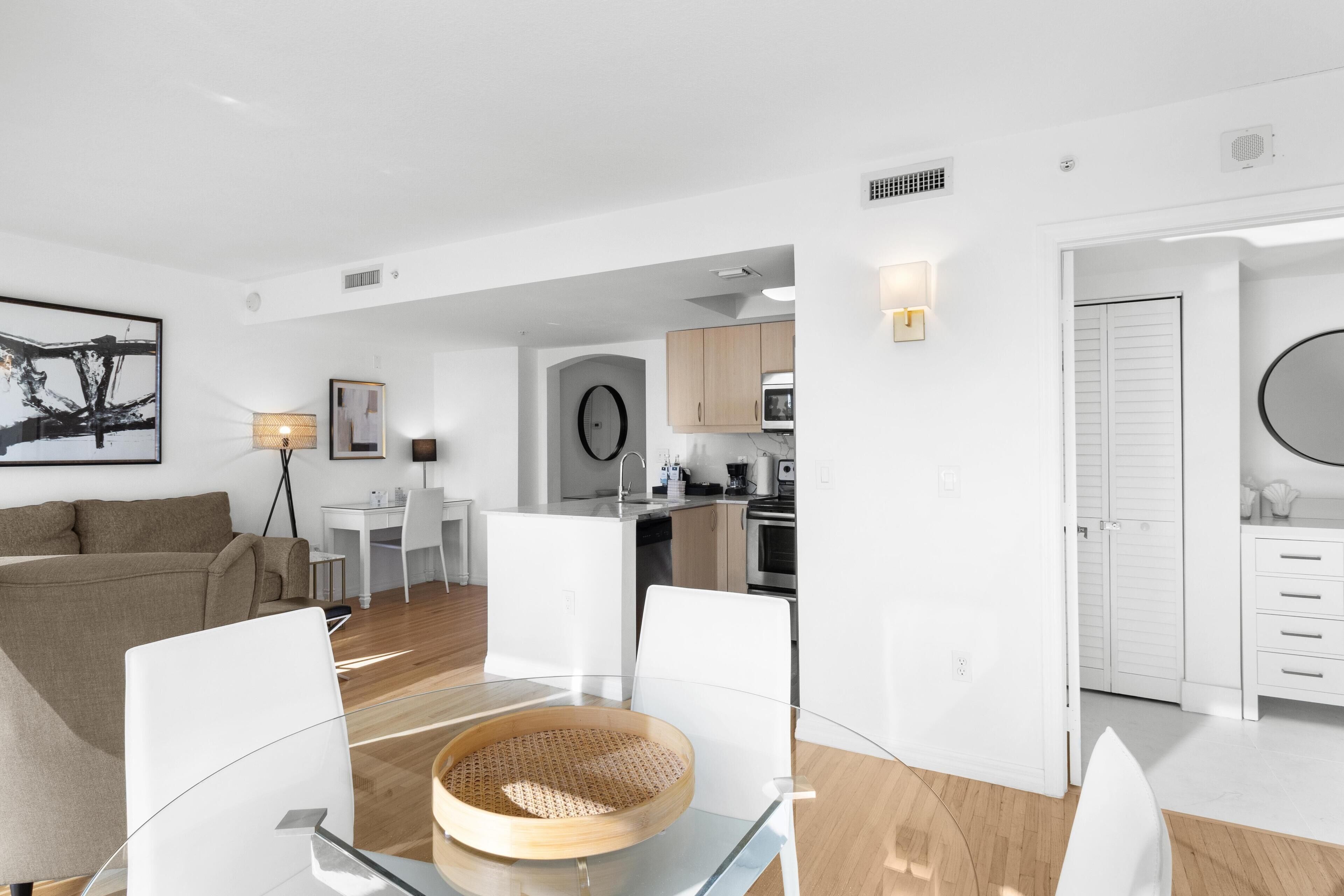 A modern living room with a glass dining table, white furniture, and a kitchen in the background.
