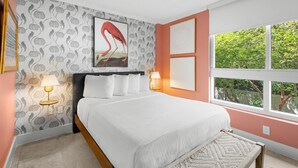 A bedroom with a bed, two bedside lamps, a window with a view of greenery, and a wall with a flamingo painting.