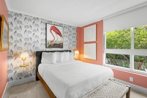 A bedroom with a bed, two bedside lamps, a window with a view of greenery, and a wall with a flamingo painting.