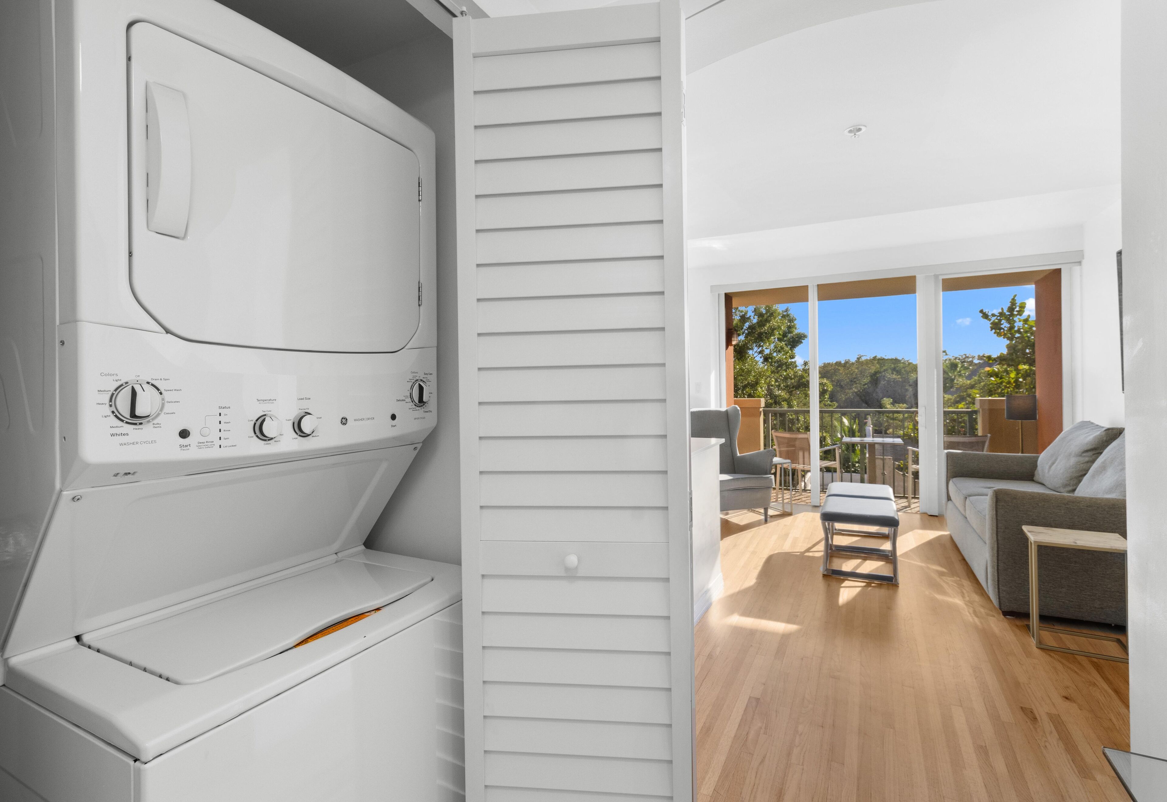 Ein moderner Waschraum mit integrierter Waschmaschine und Trockner, einer Schiebetür zu einem Balkon mit Gartenmöbeln und Blick ins Grüne.