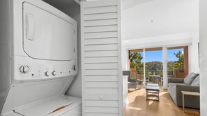 A modern laundry room with a built-in washer and dryer, a sliding door leading to a balcony with outdoor furniture and a view of greenery. - 1 Bedroom Condo: Pool & Bayfront Access (Miami)