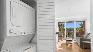 A modern laundry room with a built-in washer and dryer, a sliding door leading to a balcony with outdoor furniture and a view of greenery.