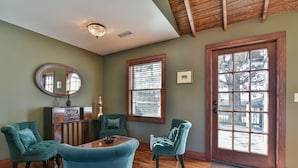Una habitación con piso de madera, un espejo grande, dos sillones tapizados en verde, una mesa de madera y una puerta con ventanal.