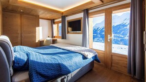 Un dormitorio con cama, techo de madera, ventanas amplias y vistas a montañas nevadas.