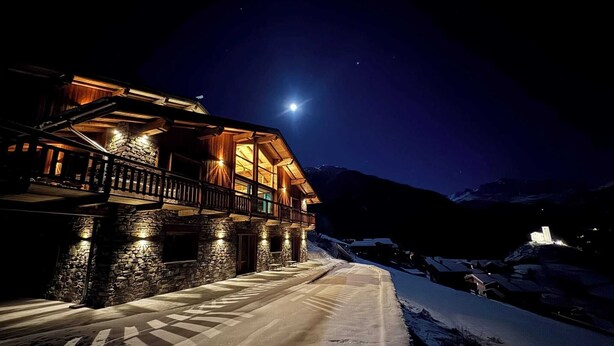 Una carretera cubierta de nieve que conduce a un edificio de madera con muros de piedra y un balcón, bajo un cielo nocturno con una luna visible.