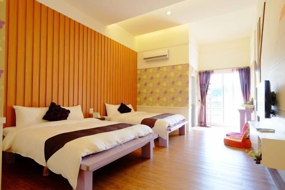 A hotel room with two beds, a wooden panel wall, an air conditioning unit, a television, and a window with curtains.