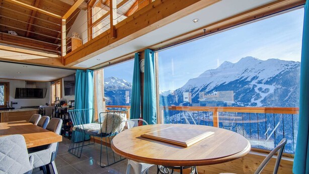 Un comedor espacioso con una mesa y sillas de madera, ventanas amplias con vistas a la montaña y un balcón con barandilla de madera.