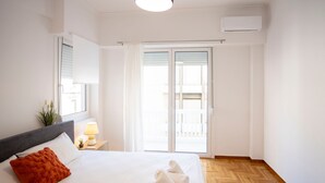 A bedroom with a bed, a nightstand, a lamp, a window with blinds, and a sliding glass door to a balcony. - Elegant 3-bedroom Retreat in the Heart of Kolonaki (Athens)